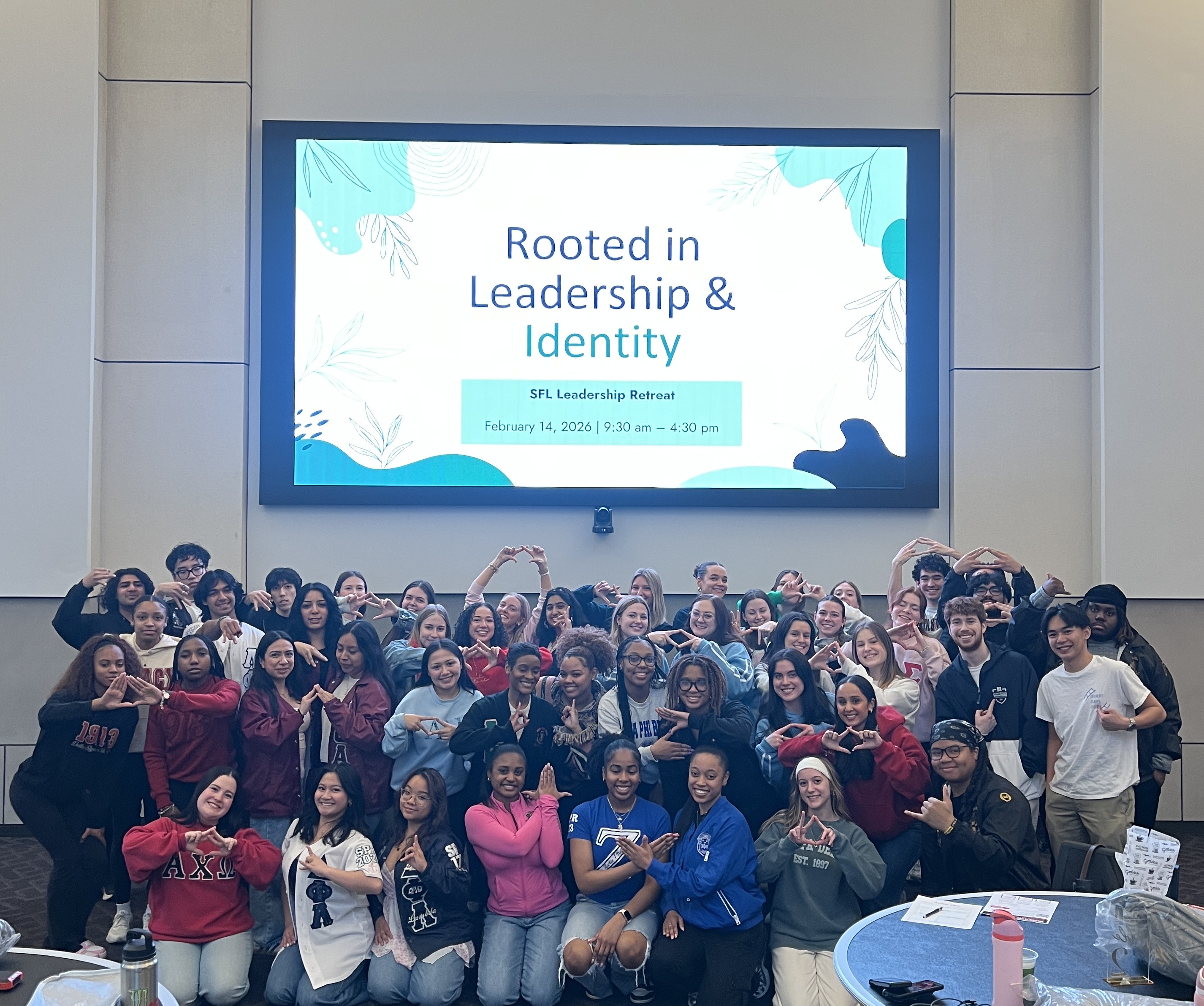 A group of students standing together in front of a wall with the mounted tv on the wall titled as Rooted in Leadership and Identity A group of students standing together in front of a wall with the mounted tv on the wall titled as Rooted in Leadership and Identity
